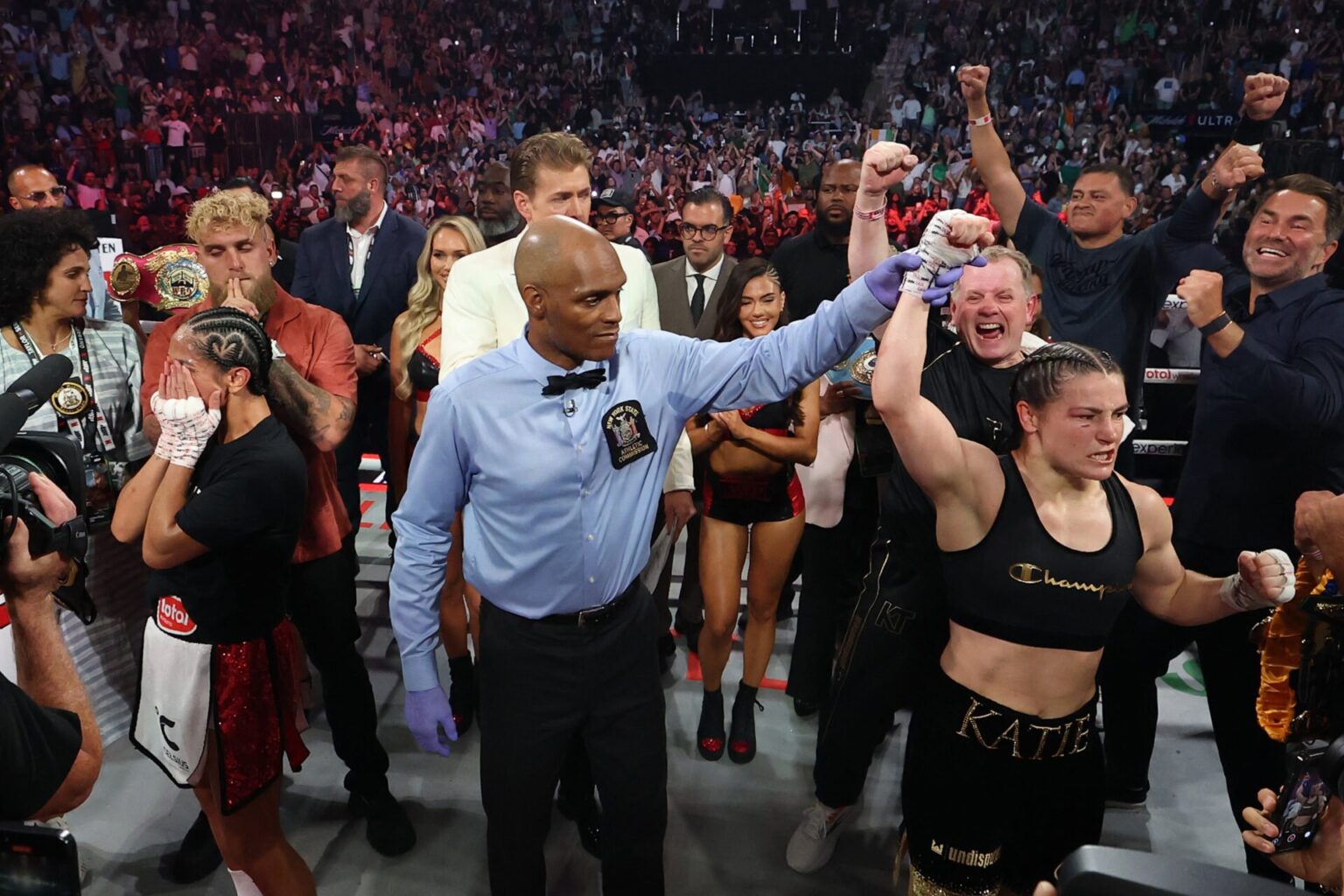 Katie Taylor has her hand raised by a referee while Amanda Serrano holds her head in her hands and a crowd gathers in the ring after their third bo...