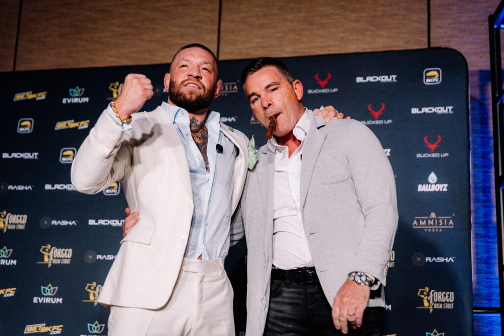Conor McGregor in a white suit does a fist pump as David Feldman smokes a cigar at the BKFC press conference