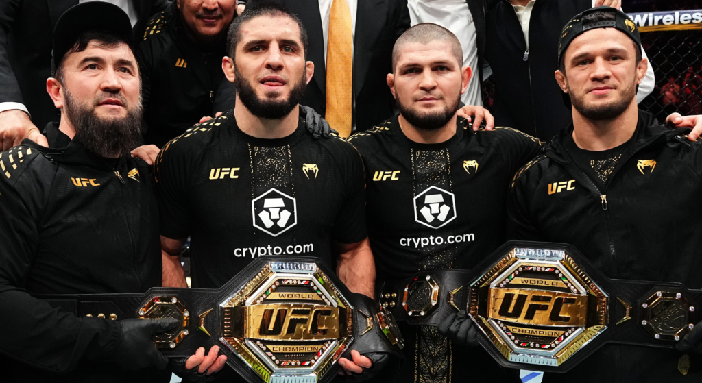Islam Makhachev and Khabib Nurmagomedov, flanked by Usman Nurmagomedov and their coach, holding Makhachev's two UFC belts