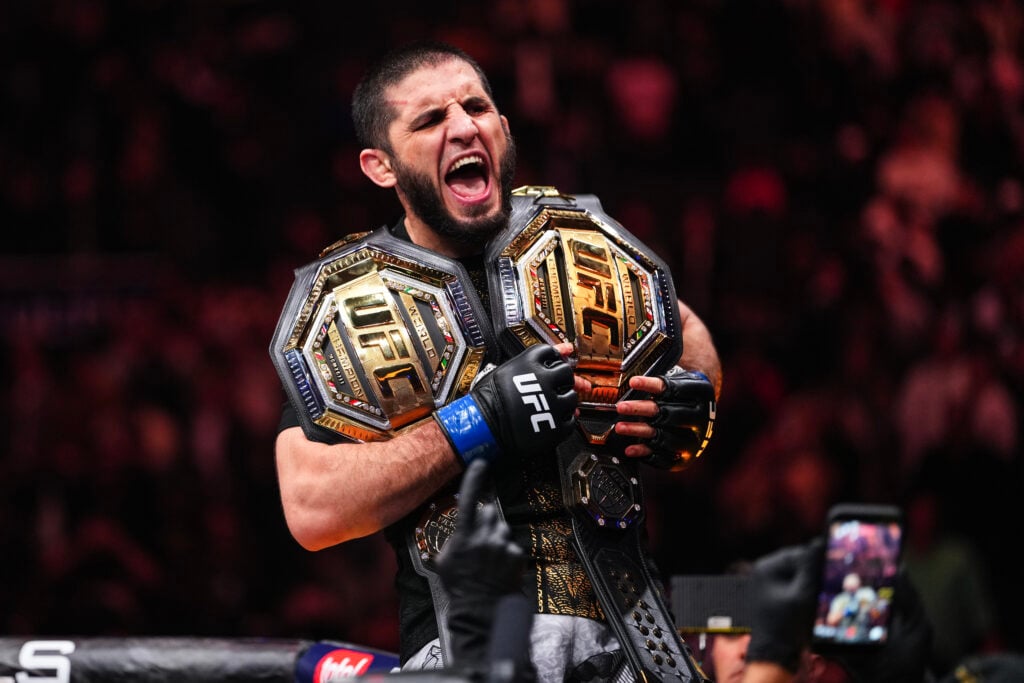 Islam Makhachev celebrates at the top of the Octagon with two UFC belts in hand